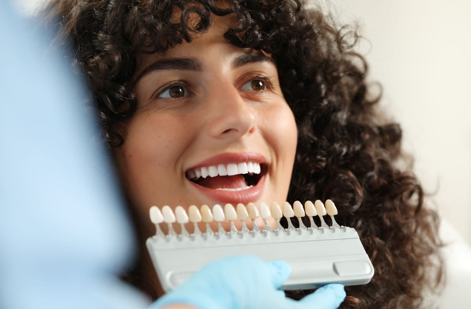 Hand in surgical glove holds up row of veneers to the mouth of a smiling woman with dark curly hair.